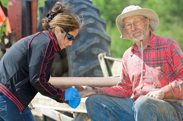 An elderly man in a straw hat and red plaid shirt is seated outdoors, while a woman in sunglasses and a black jacket is checking his blood sugar. She is wearing blue gloves and holding a small device to his finger. A tractor tire is visible in the background, indicating a rural or farm setting.