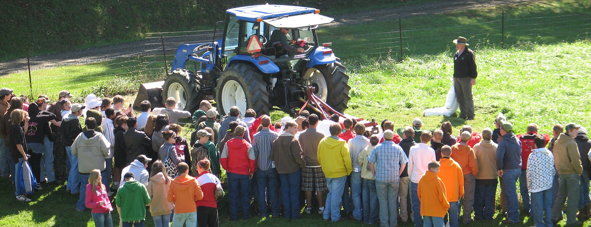 A large group of people, including teenagers and adults, are gathered in a grassy area around a blue tractor. An individual, possibly the instructor, stands to the side of the tractor, leading the session. The setting is outdoors near a wooded area. The group seems to be participating in an educational or demonstration event on agriculture.