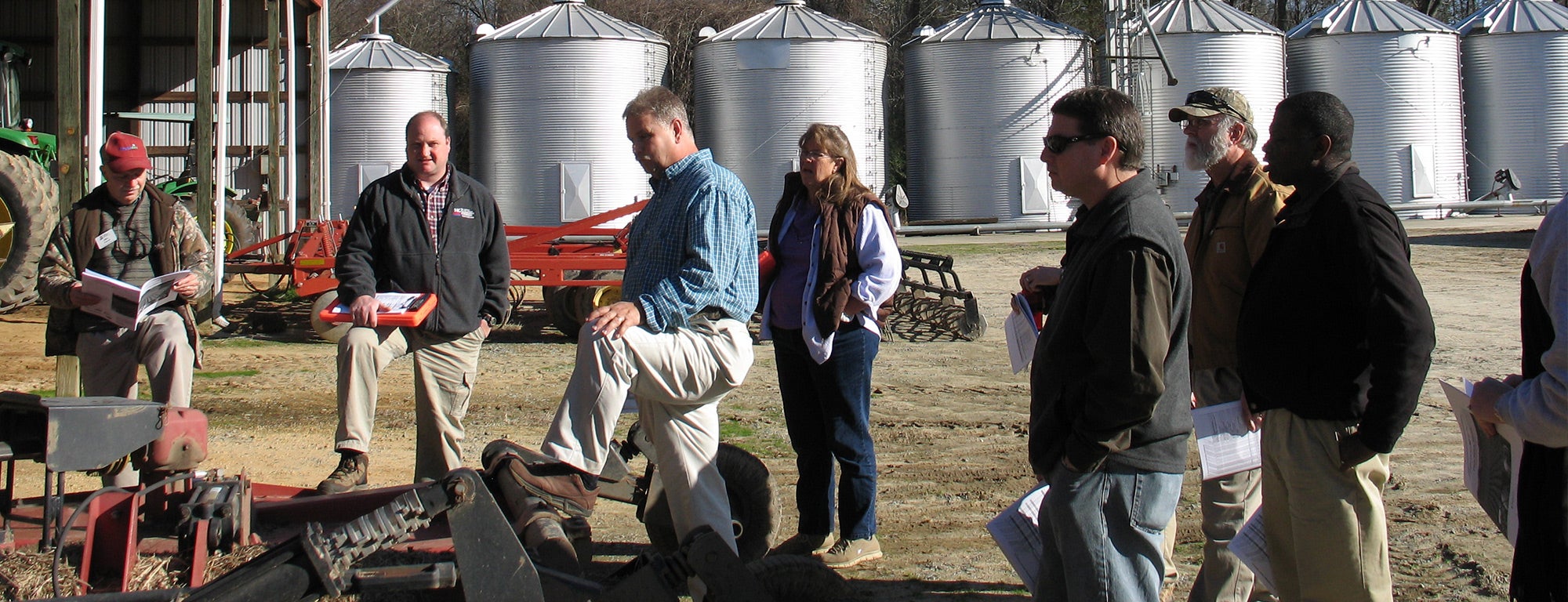 A group of people are gathered outside near a large agricultural facility. Several grain silos and structures are in the background. One man, with his leg raised and resting on a piece of machinery, appears to be leading a discussion or training session. Some individuals are holding papers and appear to be engaged in a discussion or training session. The sky is clear and blue, indicating a sunny day.