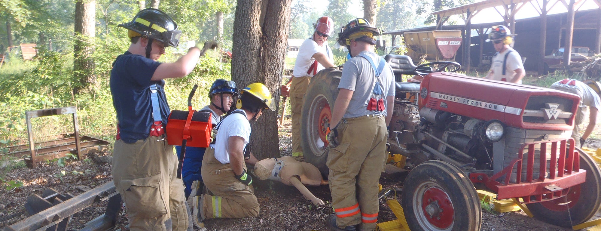 A group of firefighters in full gear are participating in a rescue training exercise. They are gathered around a red Massey Ferguson tractor, which is positioned near a tree. One firefighter is holding a bright yellow stretcher, while others are working with rescue equipment around the tractor. The scene takes place outdoors, surrounded by trees and farm structures.