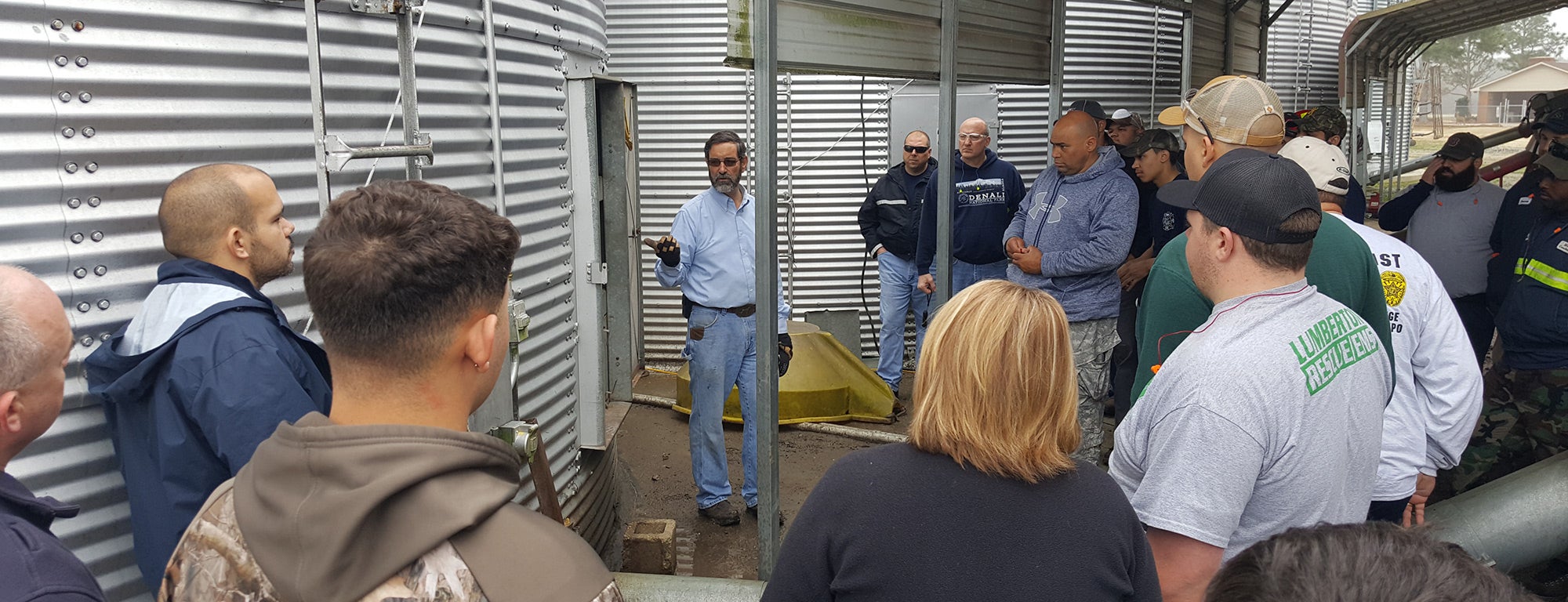 A group of people are gathered around a man who is giving a presentation near a large metal grain bin. The presenter is gesturing with one hand while the audience listens attentively. The setting appears to be an agricultural facility, with metal structures and equipment visible around the area."