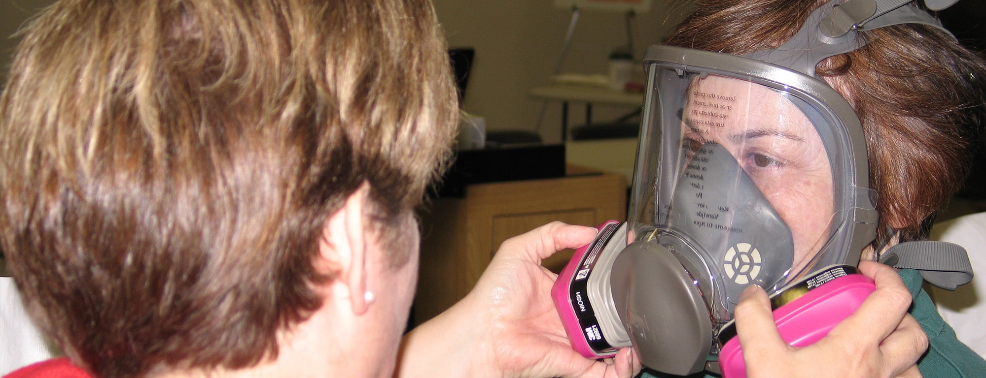 A person is being fitted with a respirator mask by another individual. The mask covers the entire face, providing a secure seal and is equipped with pink filters on either side. The fitting process appears to be taking place in a professional setting, likely for safety training or health precautions.